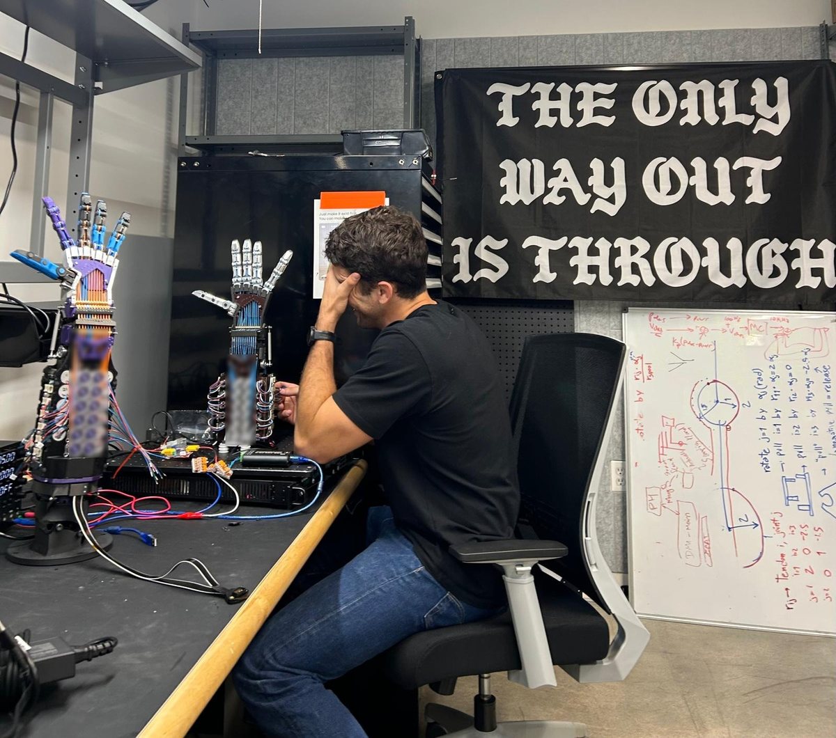 Andrea at the bench, head in hand, next to two prototype hands and a banner reading “The only way out is through”